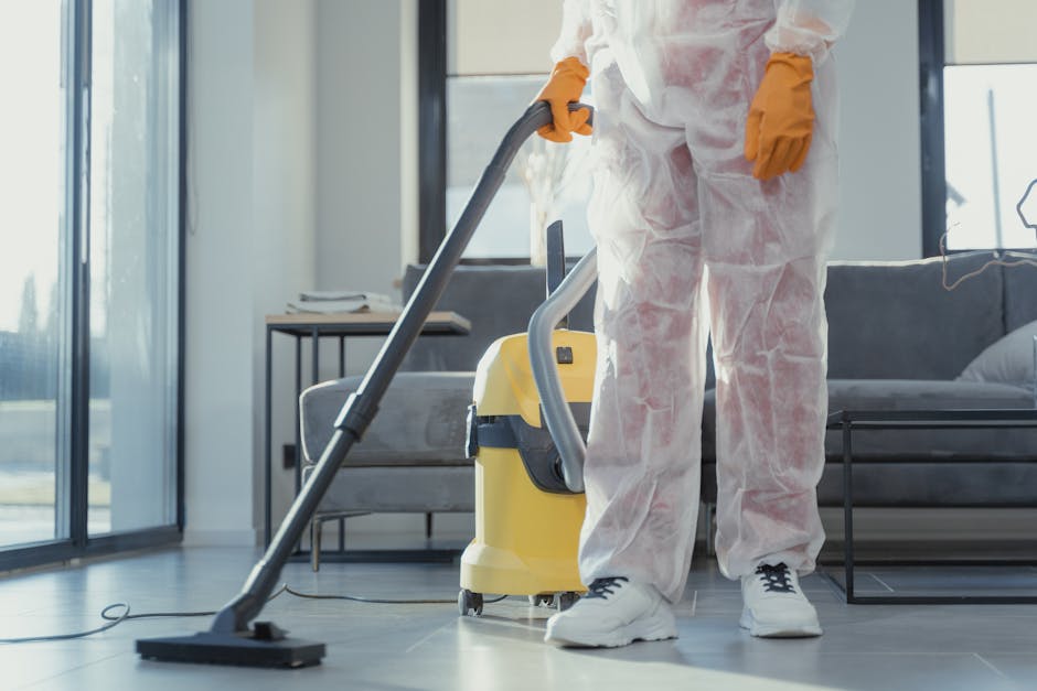 A woman wearing a yellow cleaning uniform and yellow gloves is holding a spray bottle and a blue microfiber cloth, standing in a well-lit living room with a beige sofa, wooden sideboard, and decorativ